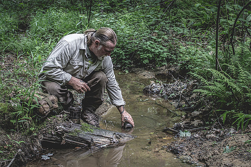 Dave Canterbury filling a bottle of water in a muddy stream