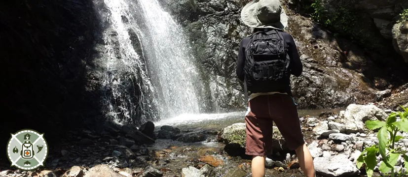 Woman with a backpack and a hat standing in front of a waterfall